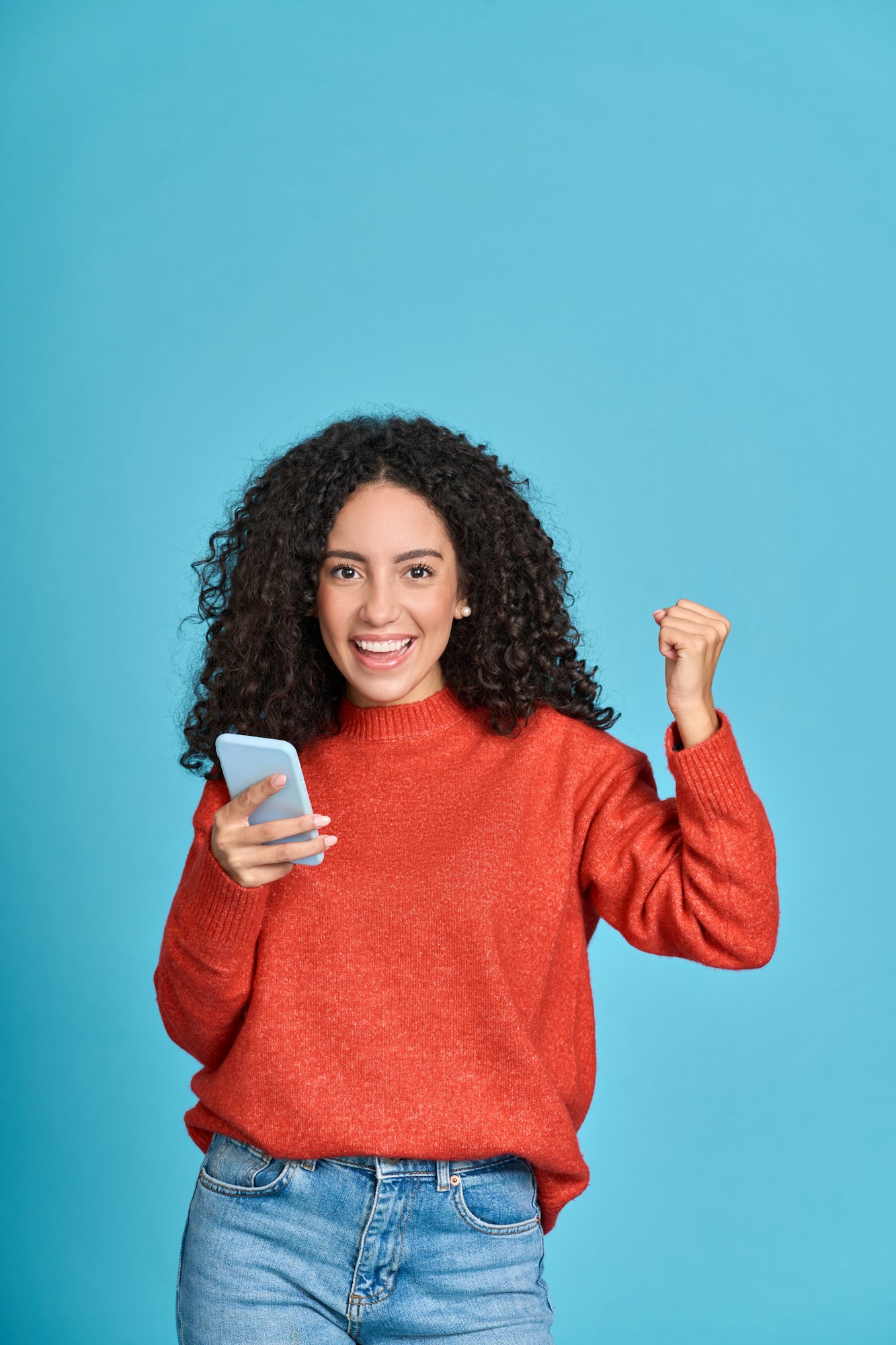 Young happy latin woman winner holding mobile cell phone isolated on blue.